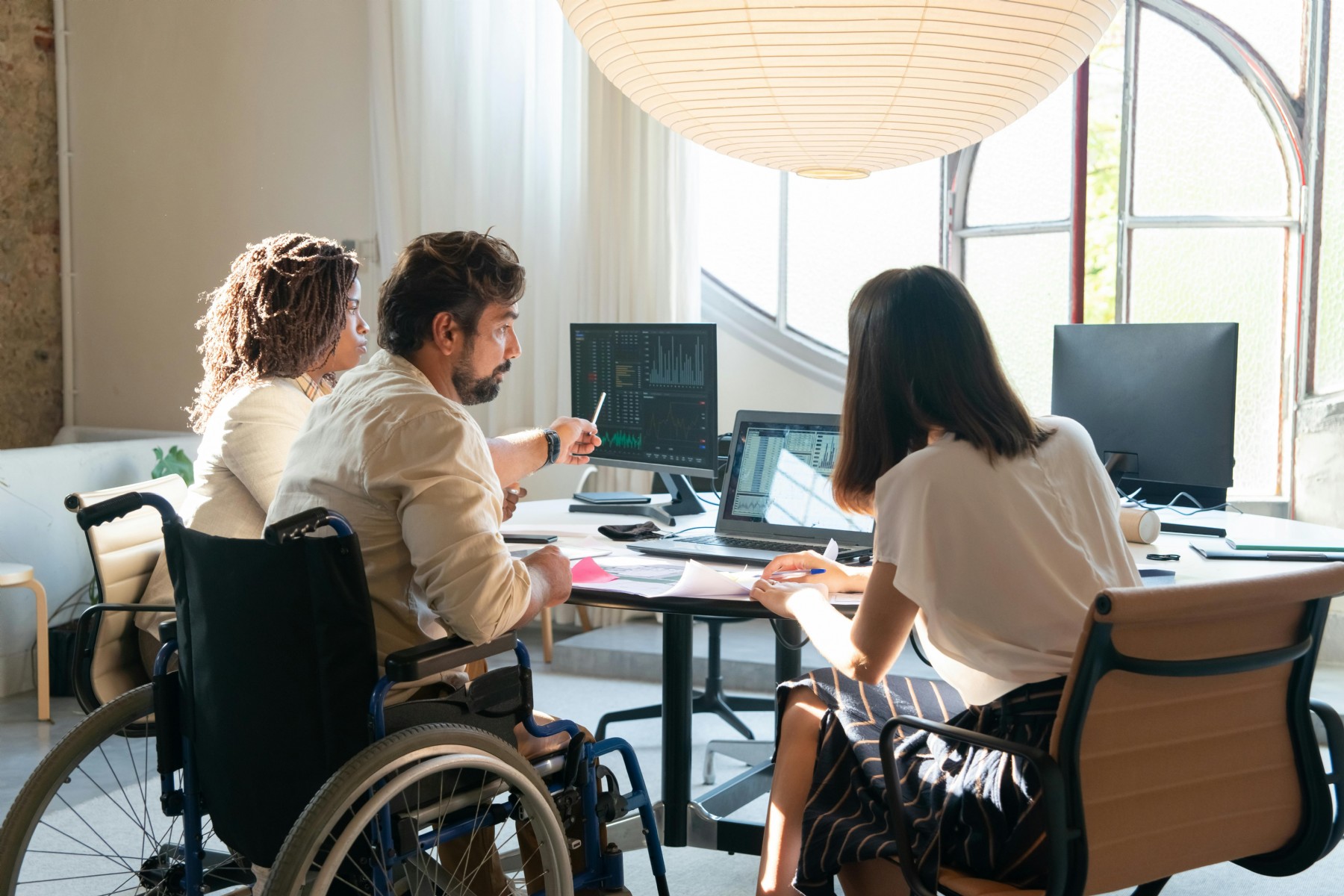 A group of individuals sitting at a table, a man in a wheelchair uses a computer