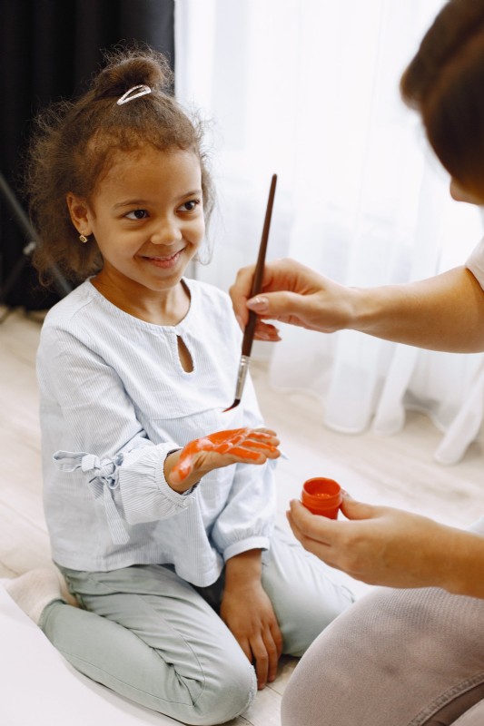 A young girl smiles while her parent holds a paintbrush, sharing a creative moment together.
