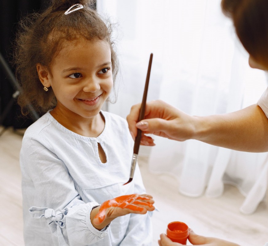 A young girl smiles while her parent holds a paintbrush, sharing a creative moment together.